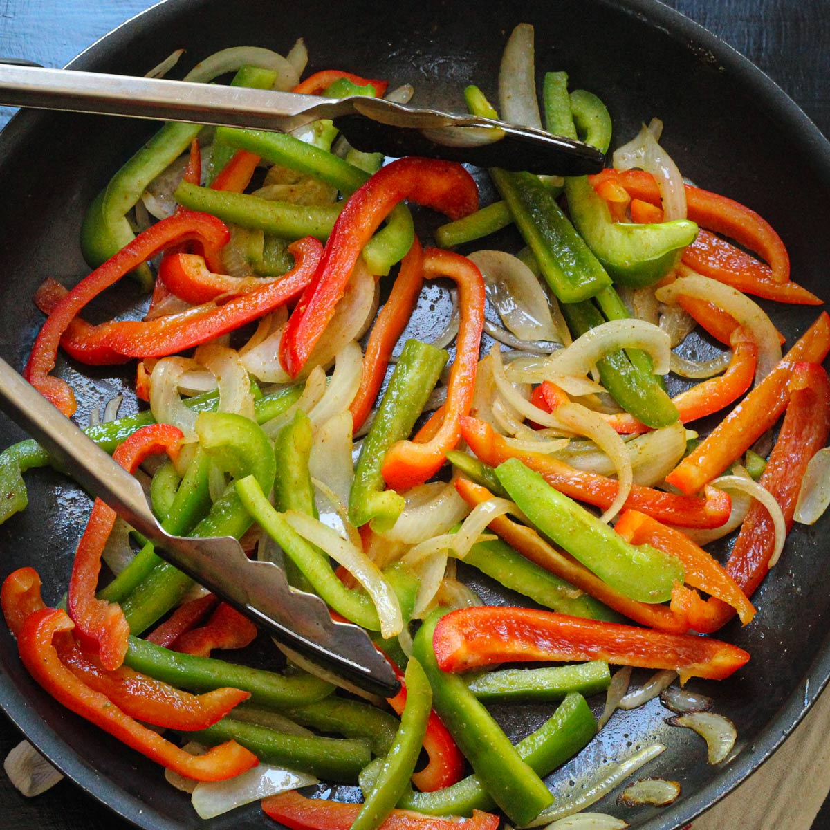 fajita vegetables in a large skillet with cooking tongs.