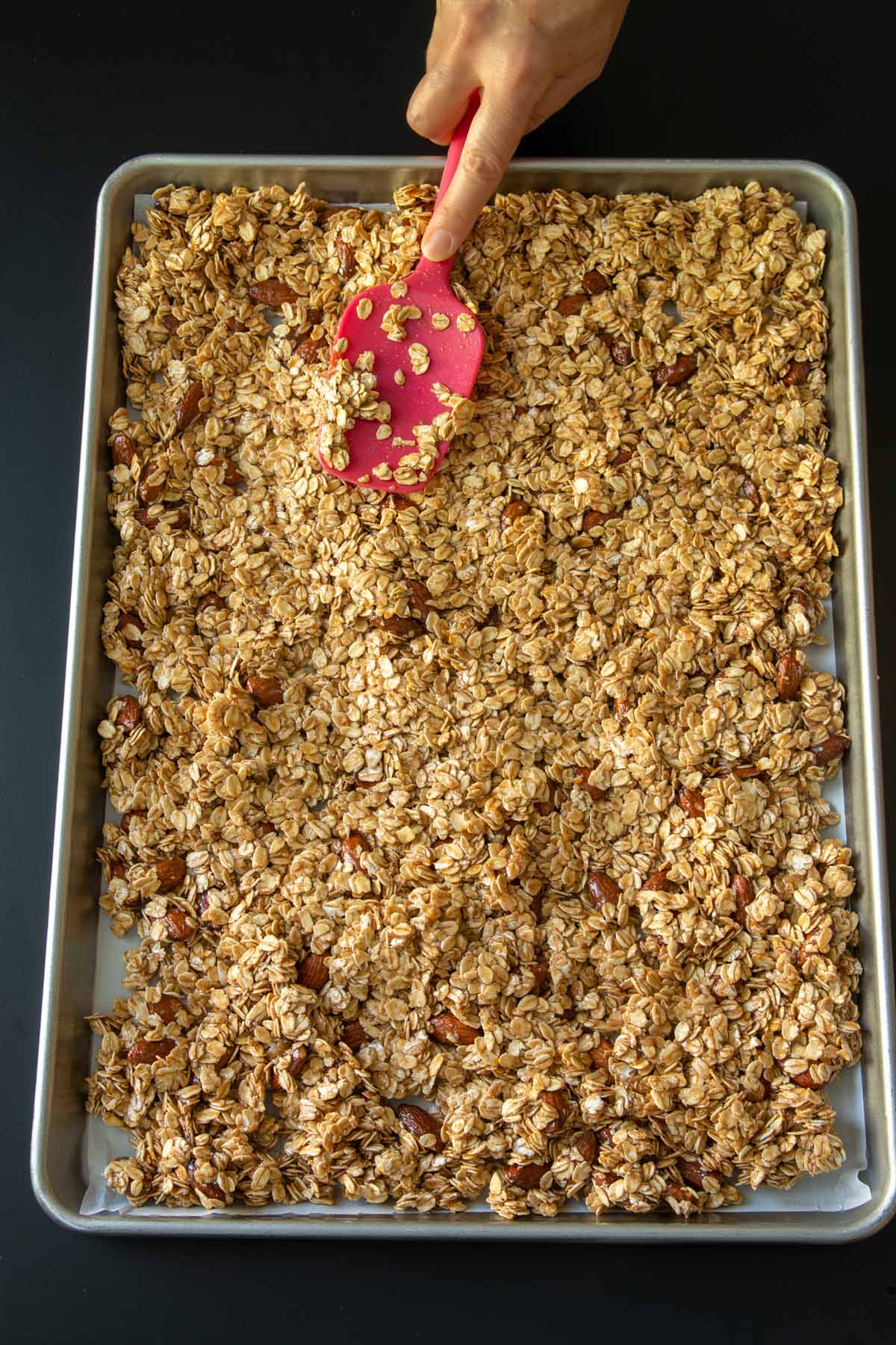 spreading granola mixture into sheet pan for baking with a red spatula.