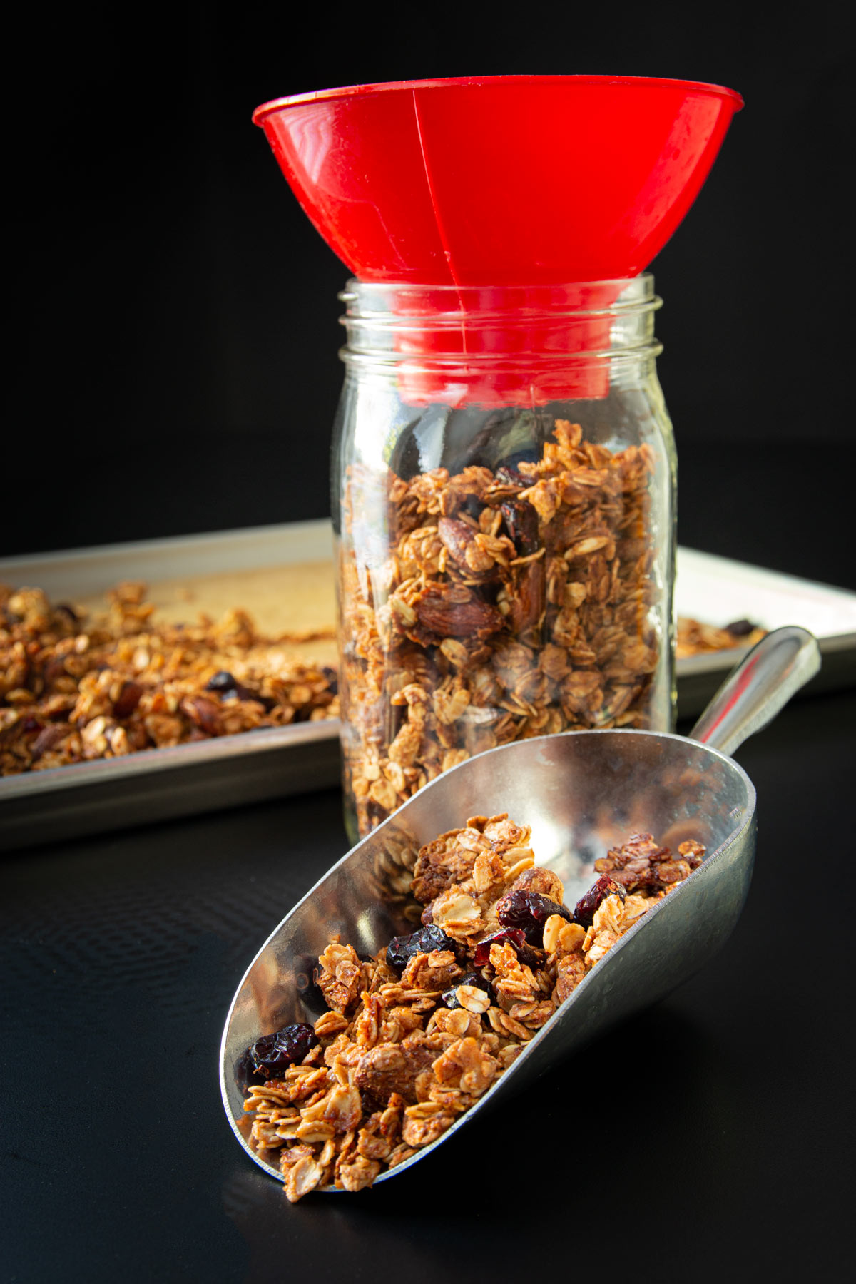 gluten free granola in pan, jar, and scoop on black table top.
