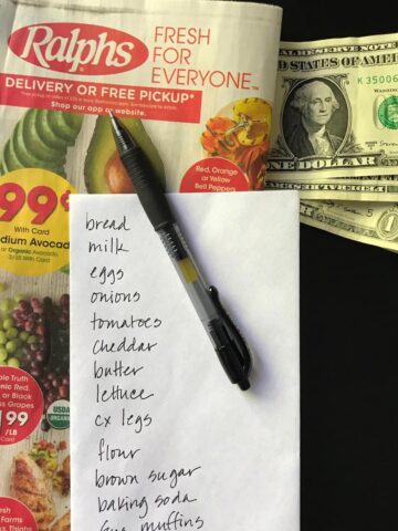 envelope grocery list atop a grocery ad with cash and a pen.