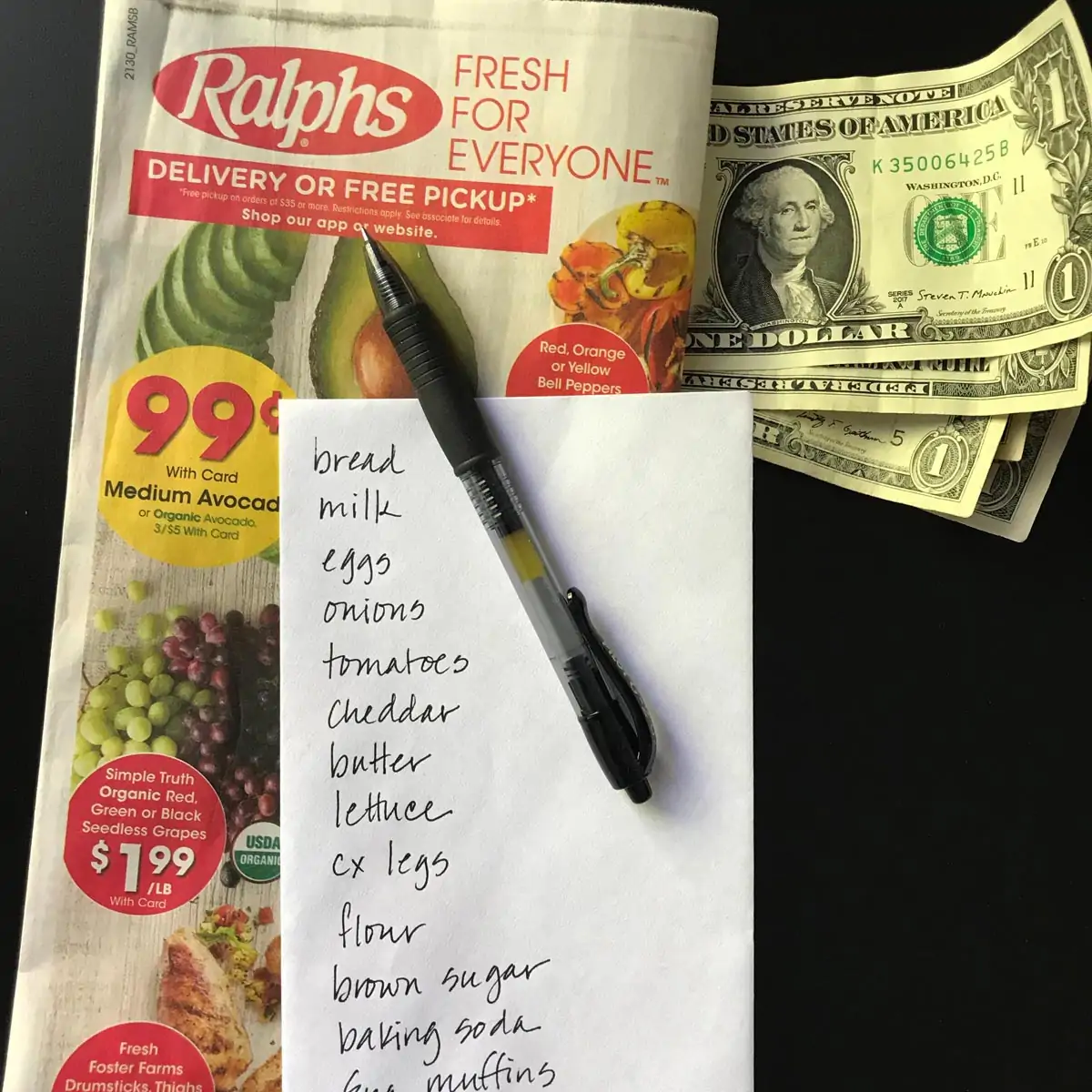 envelope grocery list atop a grocery ad with cash and a pen.