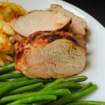 grilled pork tenderloin sliced on a plate with cheesy potatoes and green beans.