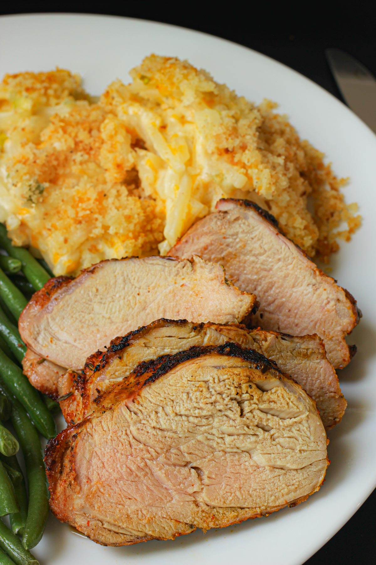 sliced grilled pork tenderloin on a plate with cheesy potatoes and green beans.