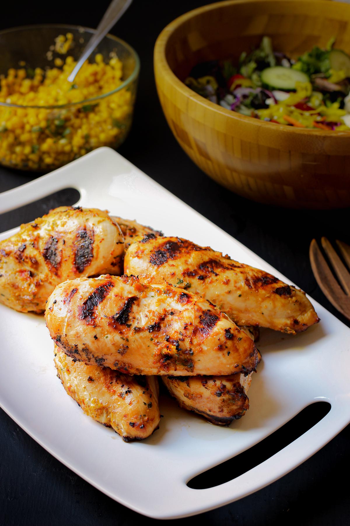 grilled spicy chicken on white platter next to salad bowl and dish of corn