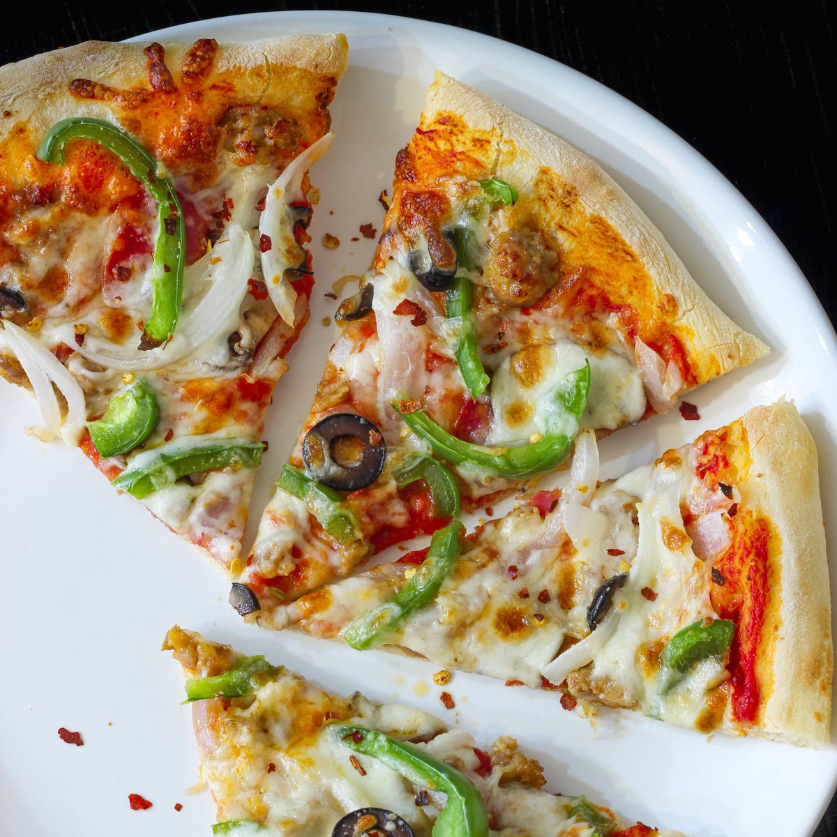 four slices of supreme pizza on a white plate on a black table top.