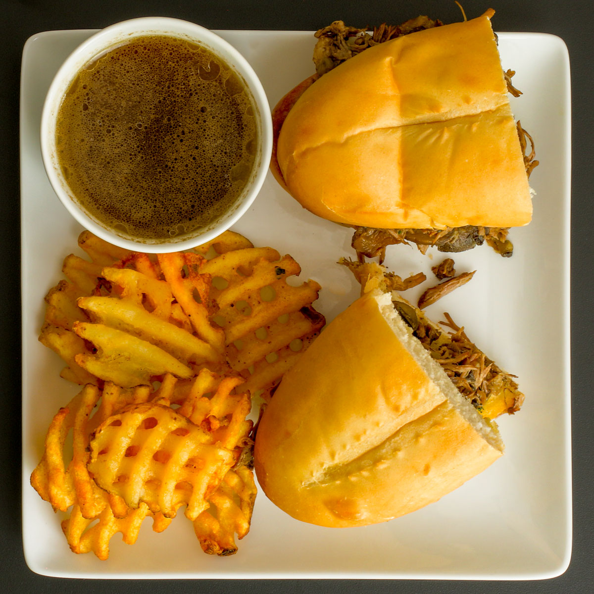 cut french dip sandwich on square plate with cup of jus and waffle fries.