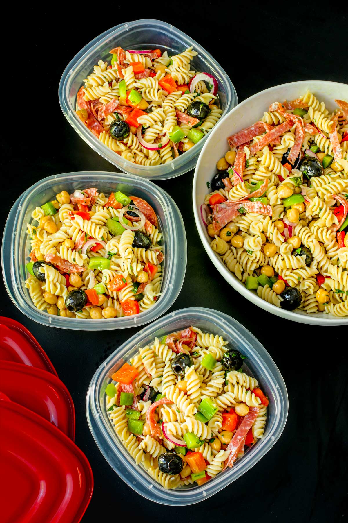 large bowl of pasta salad next to filled meal prep boxes with red lids on black table top.