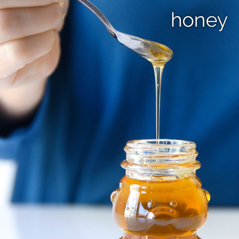 hand holding spoon dripping honey over glass bear jar.