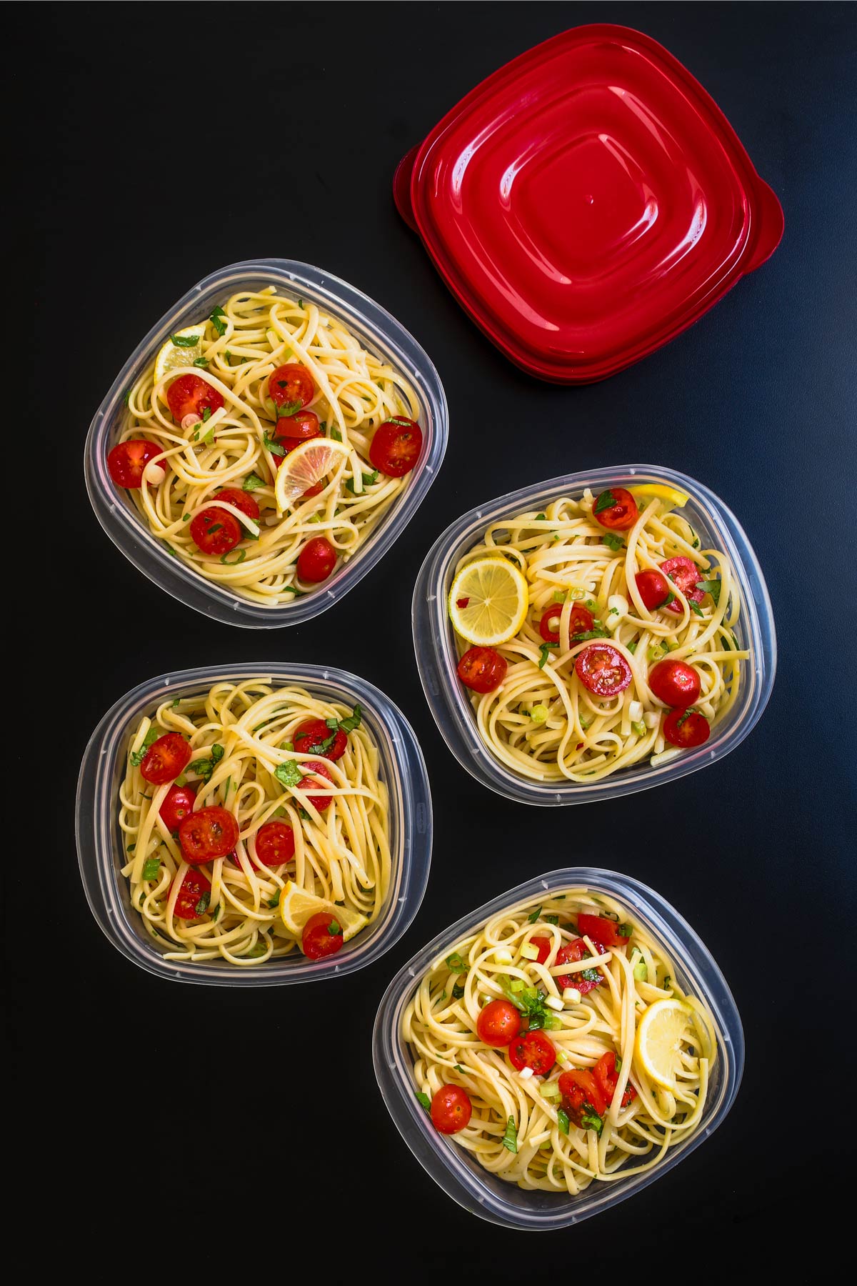 pasta with lemon divided into meal prep dishes with red lids on black tabletop.