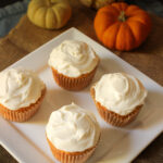 square white plate holding four frosted cupcakes next to pile of mini pumpkins.
