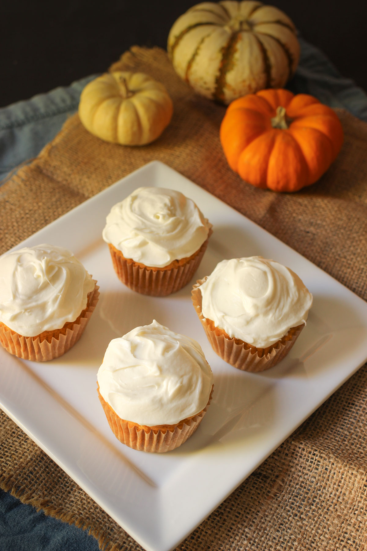 four frosted cupcakes on a square plate next to mini pumpkins.