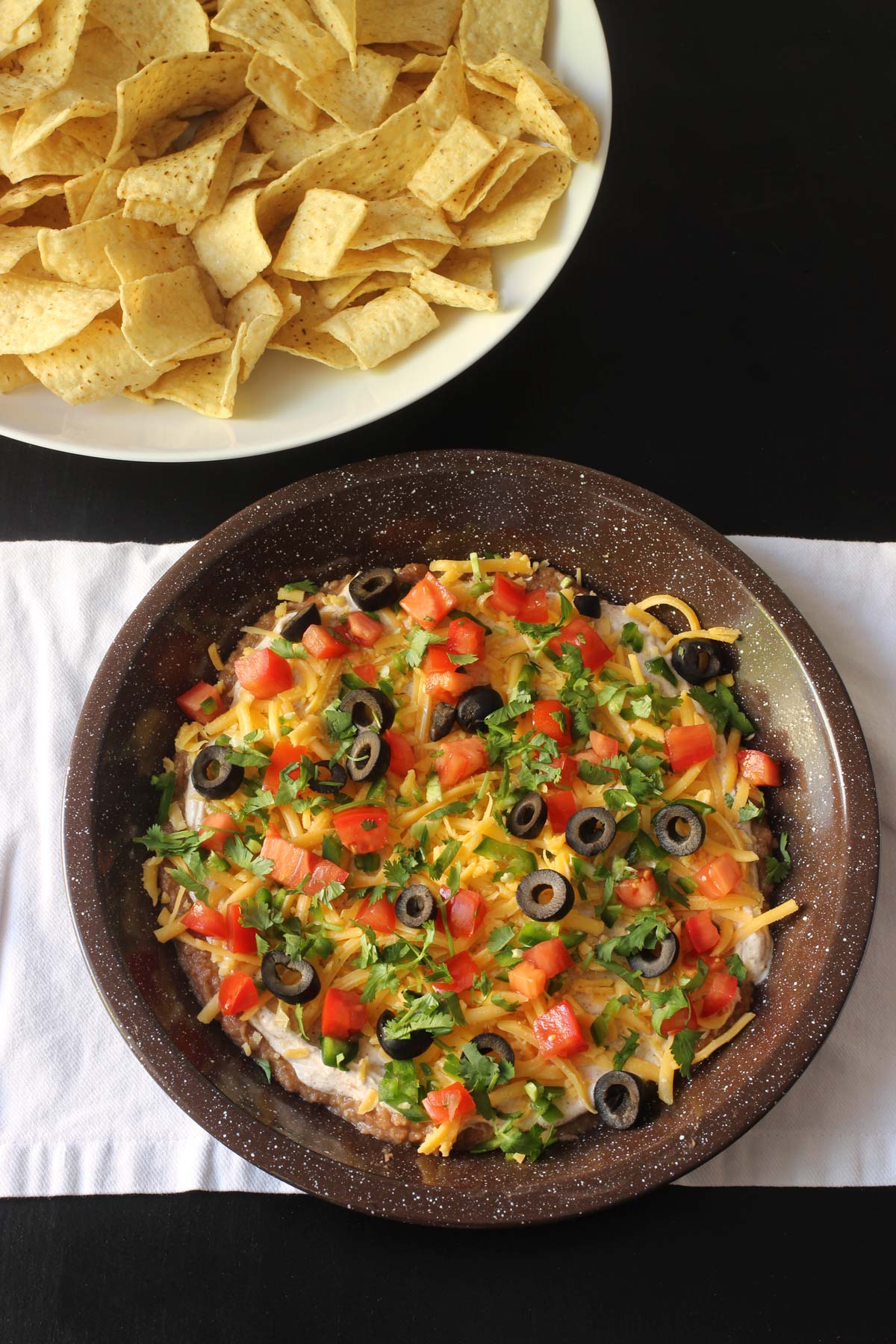 seven layer dip on white cloth next to bowl of chips.
