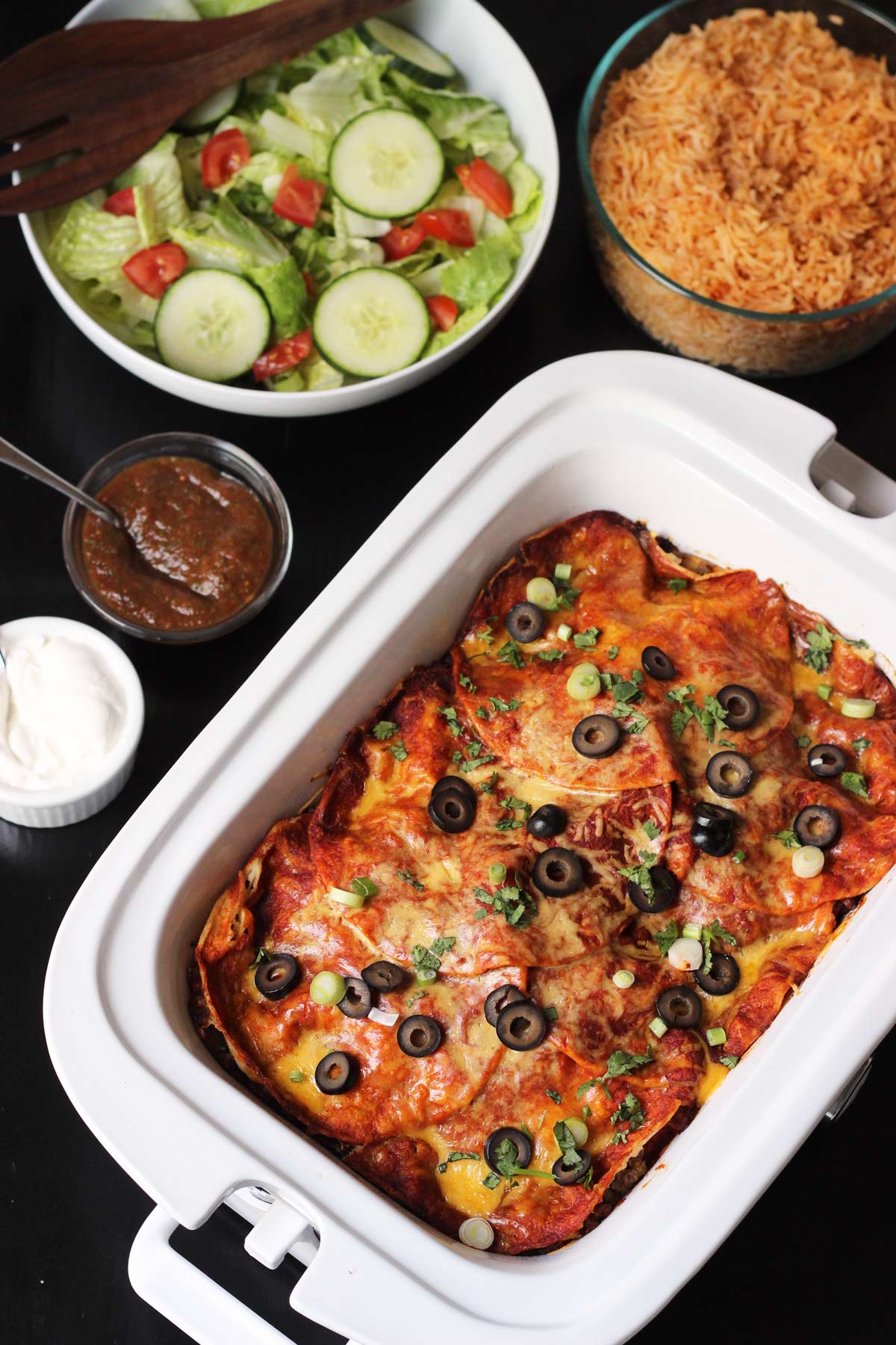 table set with casserole crock pot of enchiladas with toppings, rice, and salad.