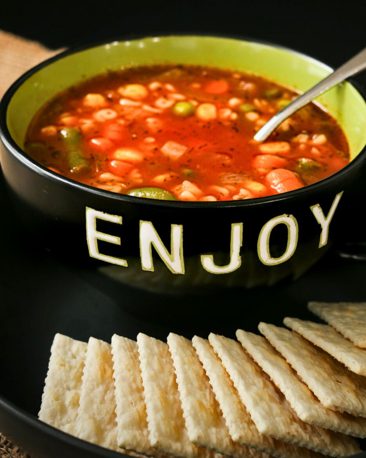 black bowl of alphabet soup on a plate with saltine crackers.