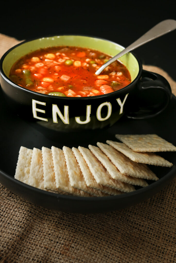 alphabet soup and a spoon in a bowl labeled enjoy on a black plate with saltine crackers.