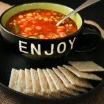 alphabet soup in black bowl marked ENJOY with plate of saltines.