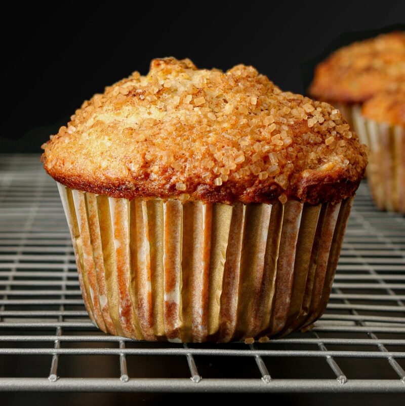 close up of apple cinnamon muffin in striped paper cooling on silver wire rack on black table.
