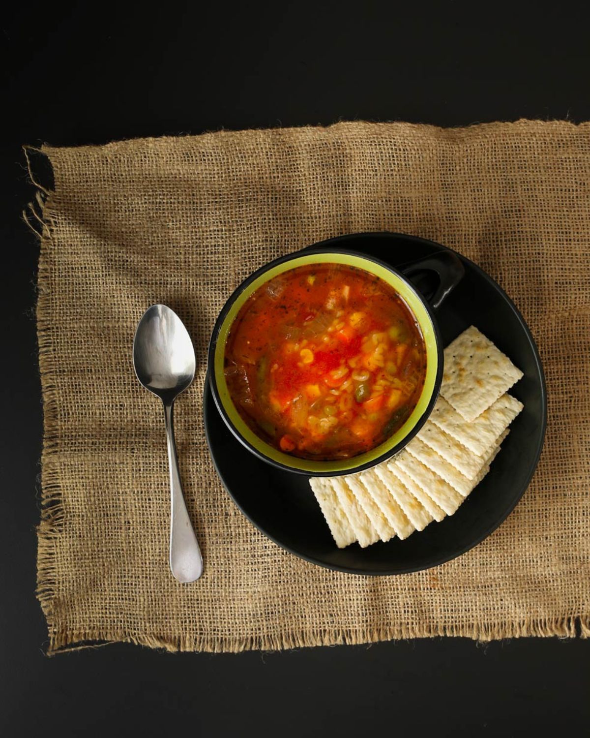black table with coarse cloth, black plate of crackers and a bowl of alphabet soup.