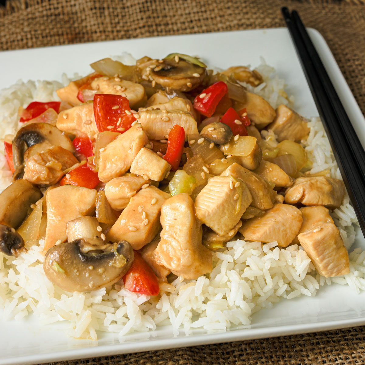 chicken stir fry on a bed of rice on a square white plate with two black chopsticks leaning on the plate.