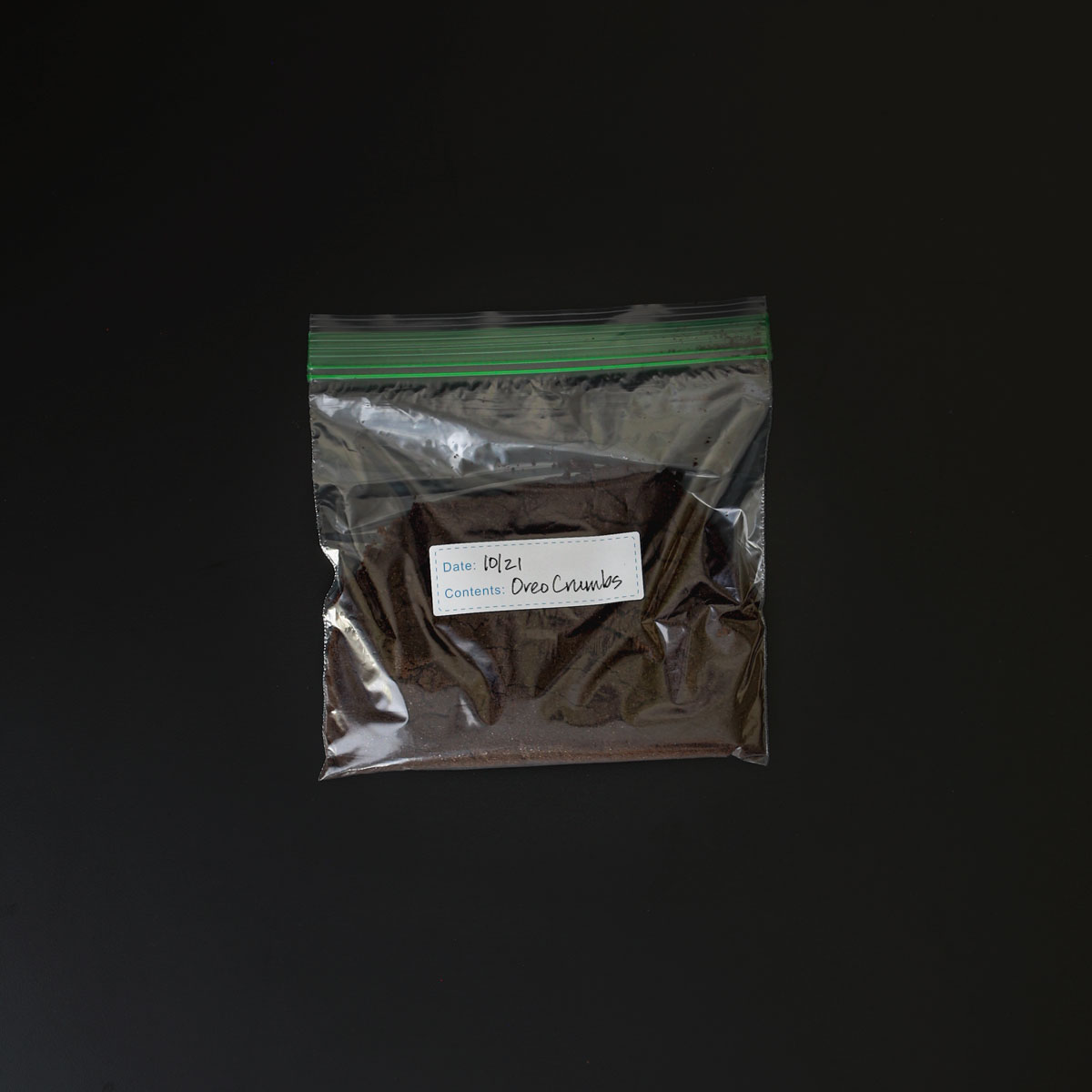 chocolate cookie crumbs stored in a labeled ziptop bag on a black table.