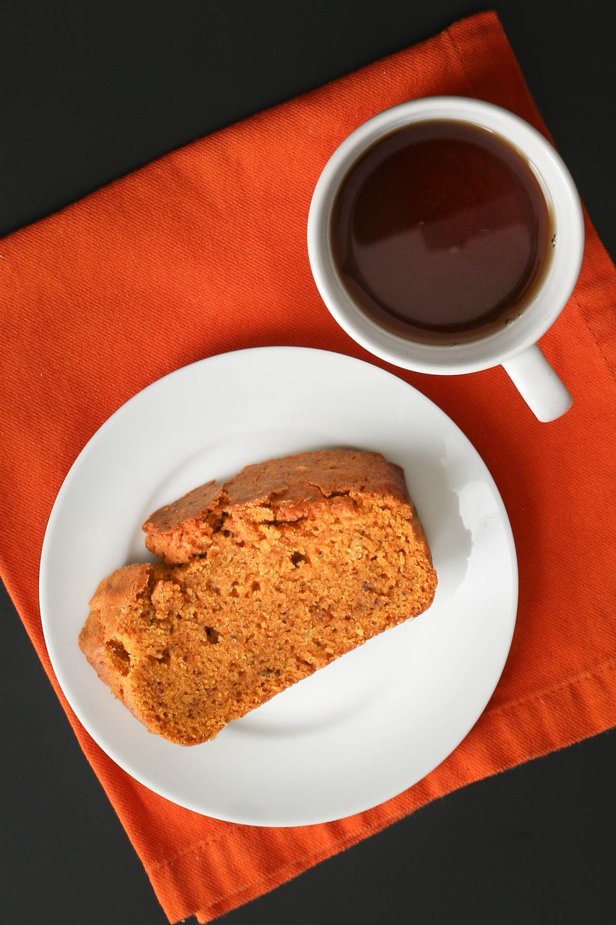 slice of vegan pumpkin bread on a white plate on an orange napkin next to a small cup of tea.
