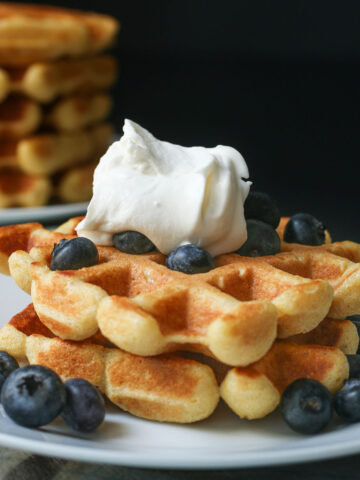 pair of cornbread waffles topped with blueberries and whipped cream on a white plate near a stack of waffles.