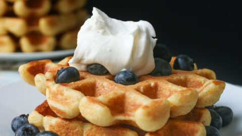 pair of cornbread waffles topped with blueberries and whipped cream on a white plate near a stack of waffles.