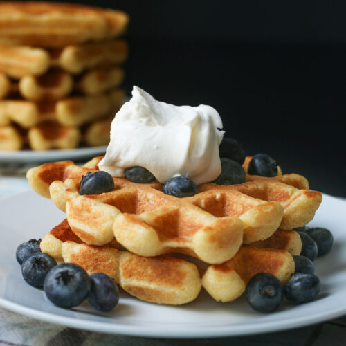 pair of cornbread waffles topped with blueberries and whipped cream on a white plate near a stack of waffles.