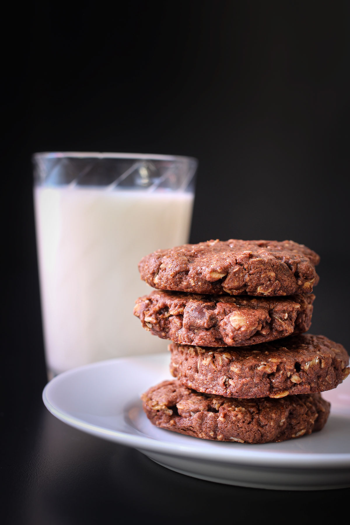 stack of chocolate oatmeal cookies on a white plate next to a glass of milk.