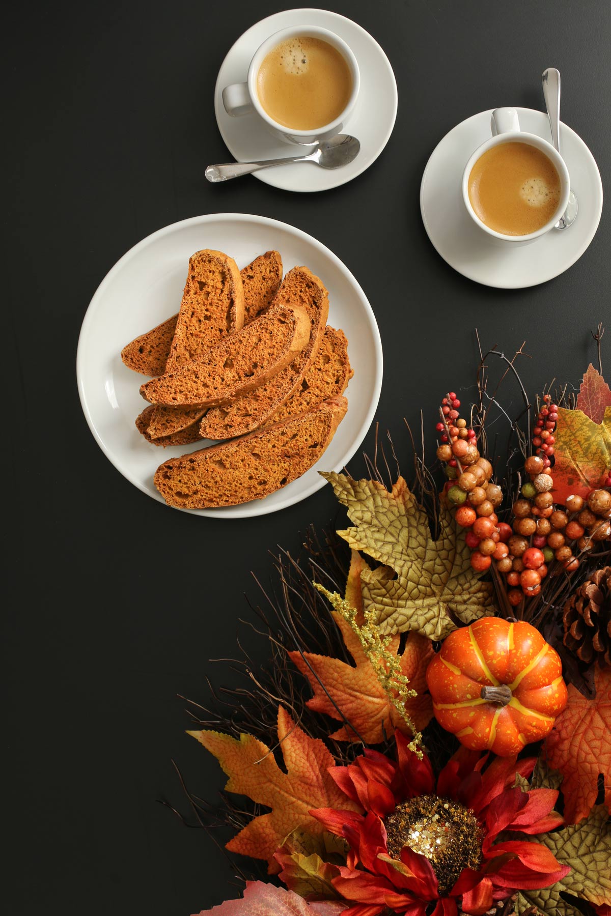cups of espresso on saucers next to platter of biscotti framed by pumpkin decor and fall leaves.