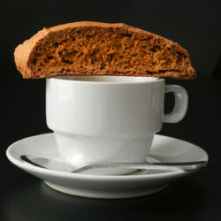 pumpkin biscotti resting on a cup of espresso on saucer with demitasse spoon.
