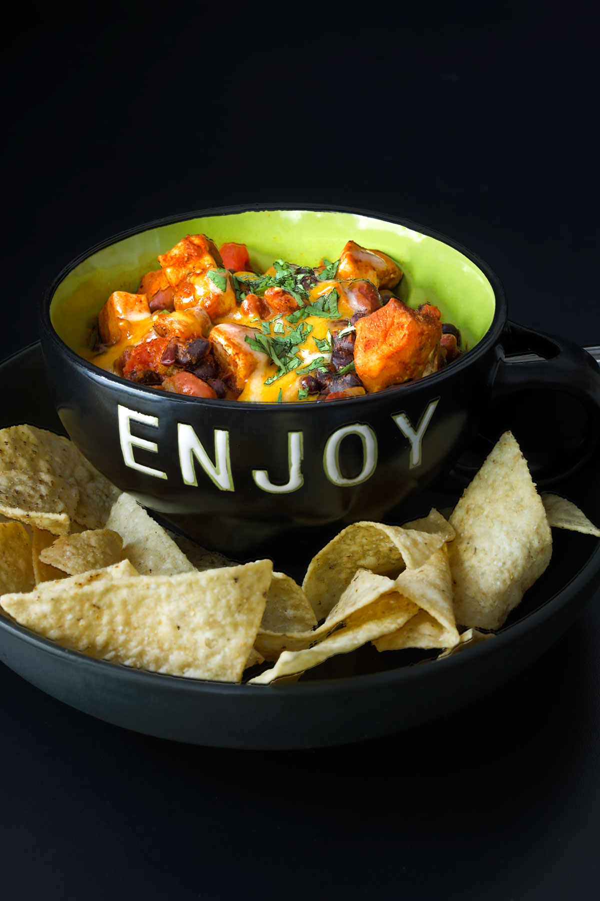 black bowl of chili marked ENJOY on plate of chips on black table top.