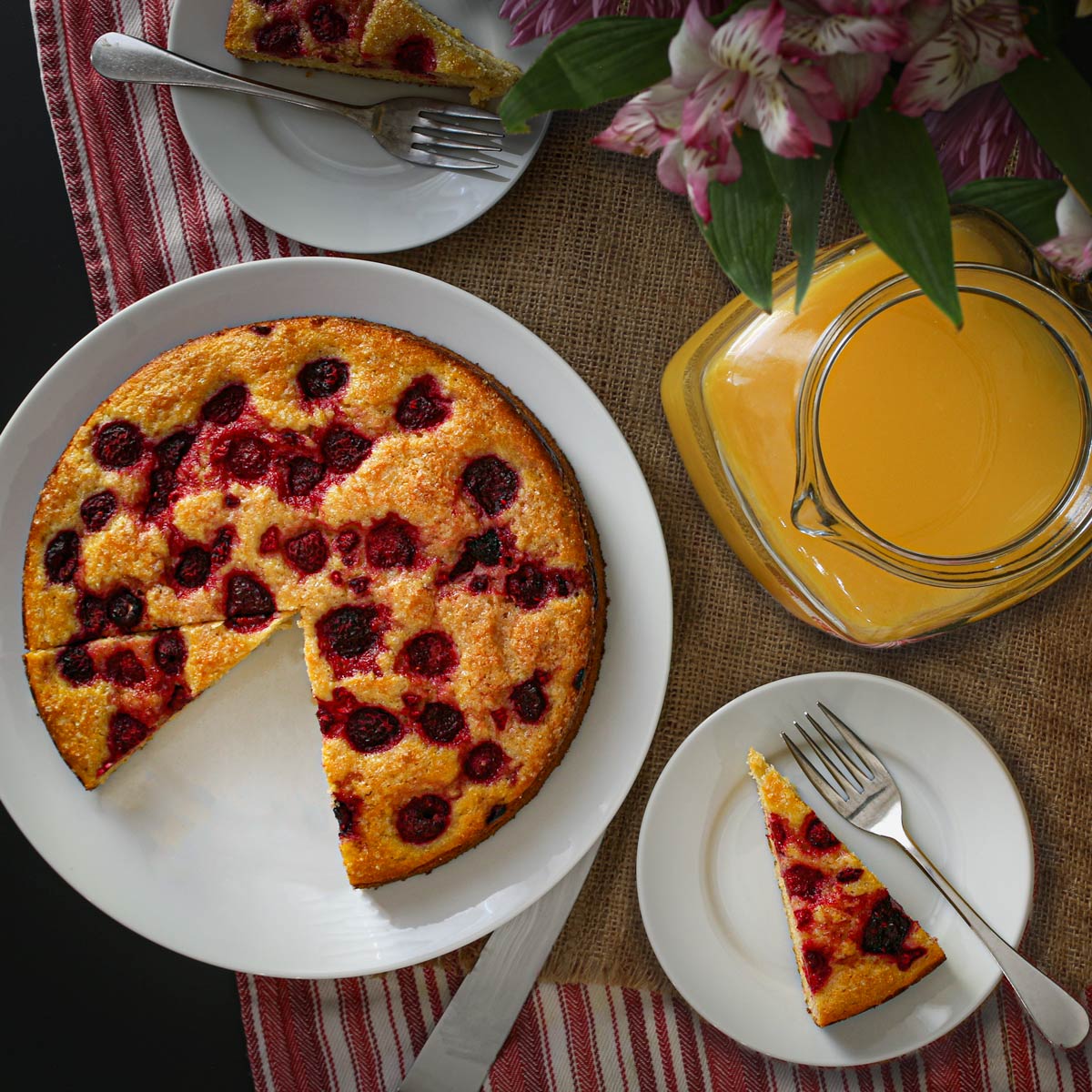 platter with breakfast cake cut into wedges next to dishes with slices on them with forks as well as a pitcher of orange juice and a vase of flowers on the table.