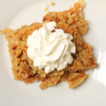 square of apple pie on white plate topped with whipped cream.