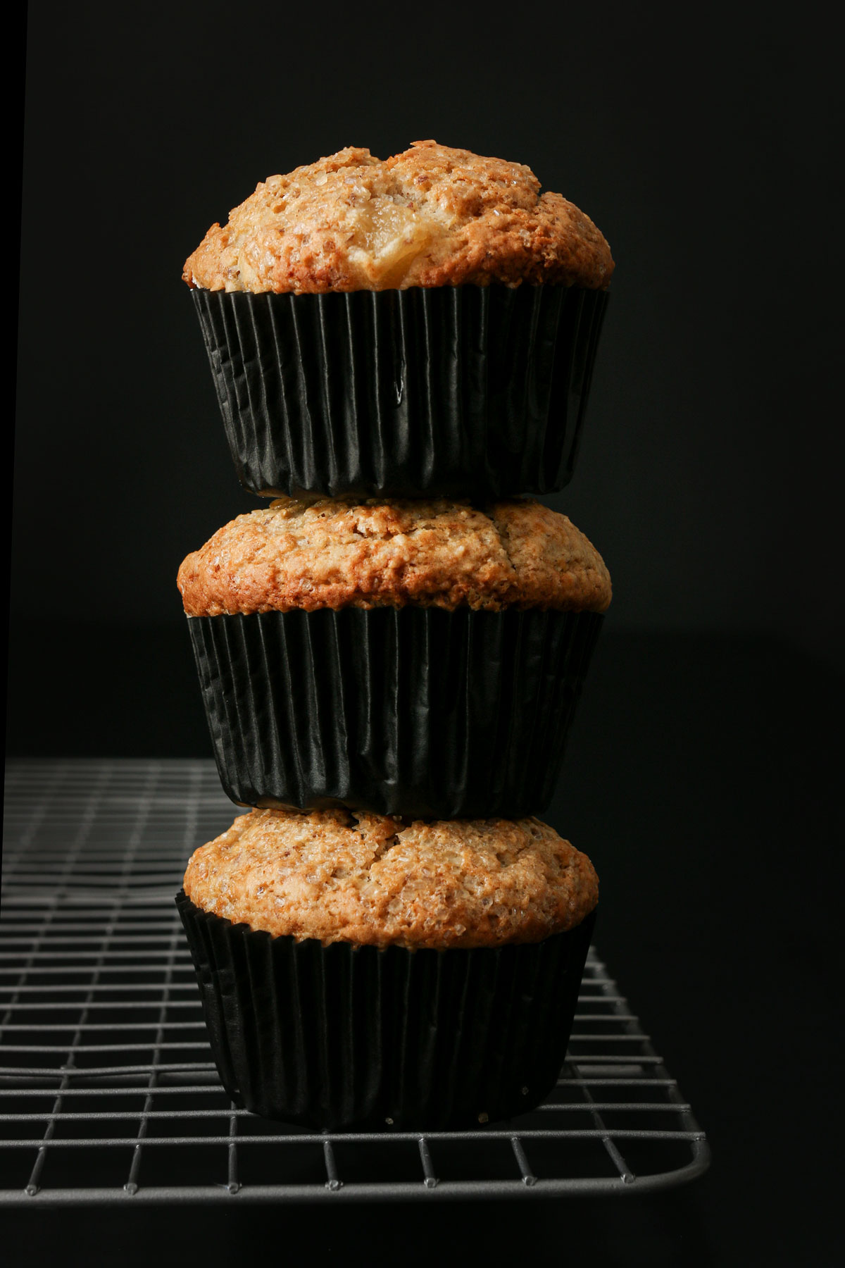 stack of three vegan muffins in black paper muffin cups. on the corner of the cooling rack on a black table.