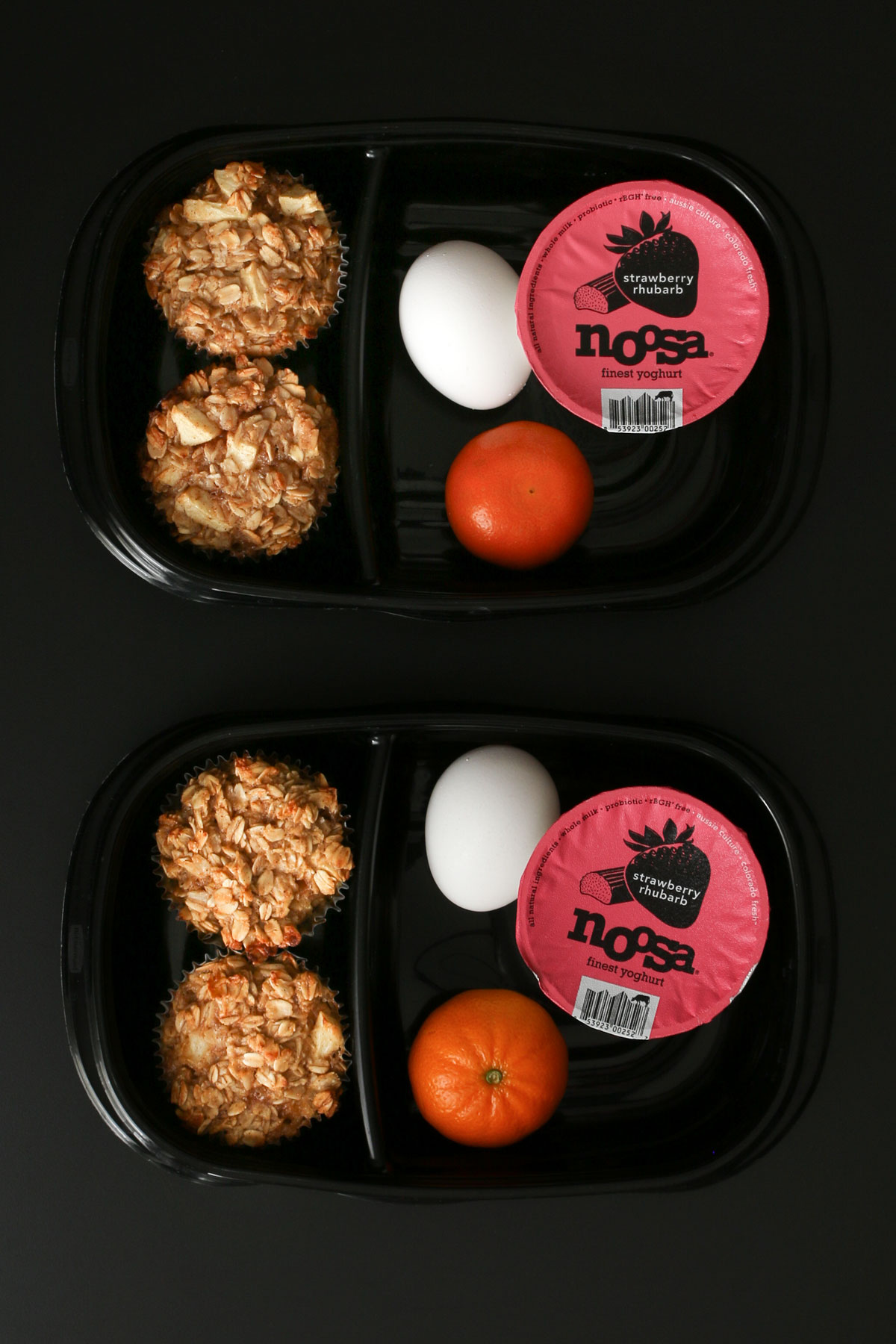 two black divided meal prep boxes each with two baked oatmeal cups in one section and hardcooked egg, clementine, and yogurt cup in the other section.