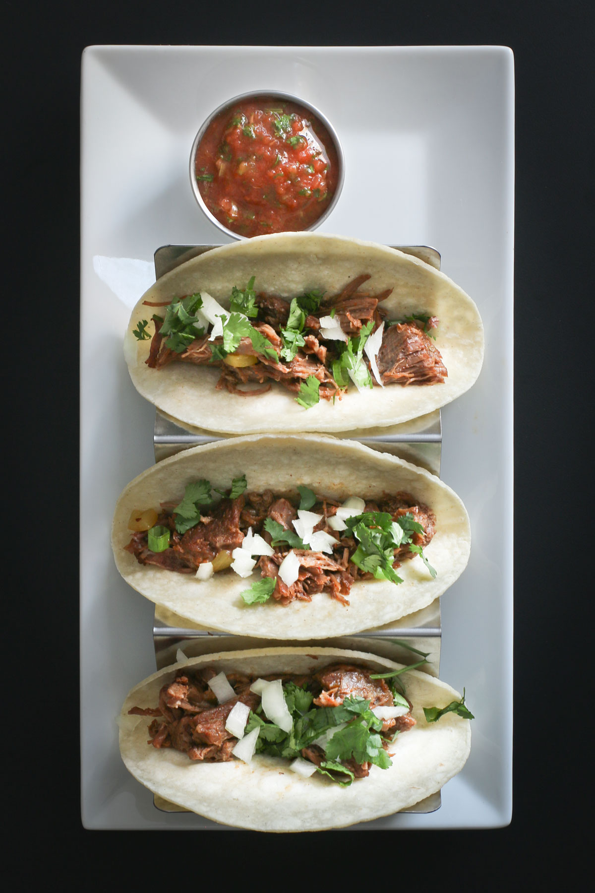 shredded beef tacos on a taco tray on a white platter with a cup of salsa.