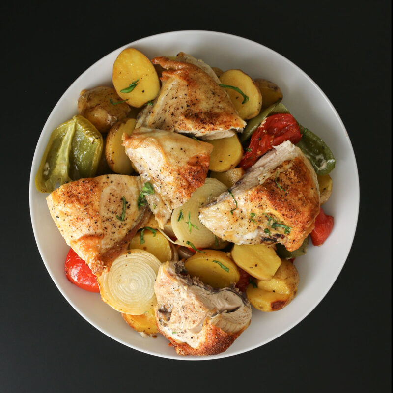 baked chicken breasts cut in halves sitting atop a platter of roasted potatoes, peppers, tomatoes, and onions on a black table top.