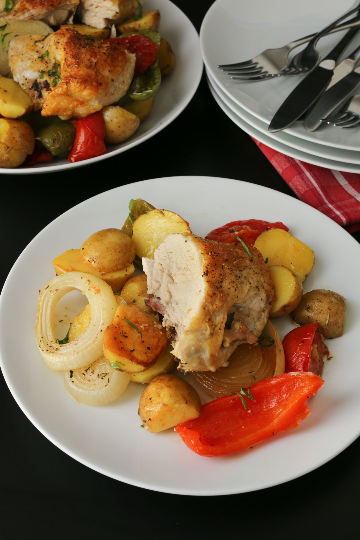 dinner plate with baked chicken atop a bed of peppers, onions, and potatoes on a dinner table set with a serving platter and additional plates and flatware.