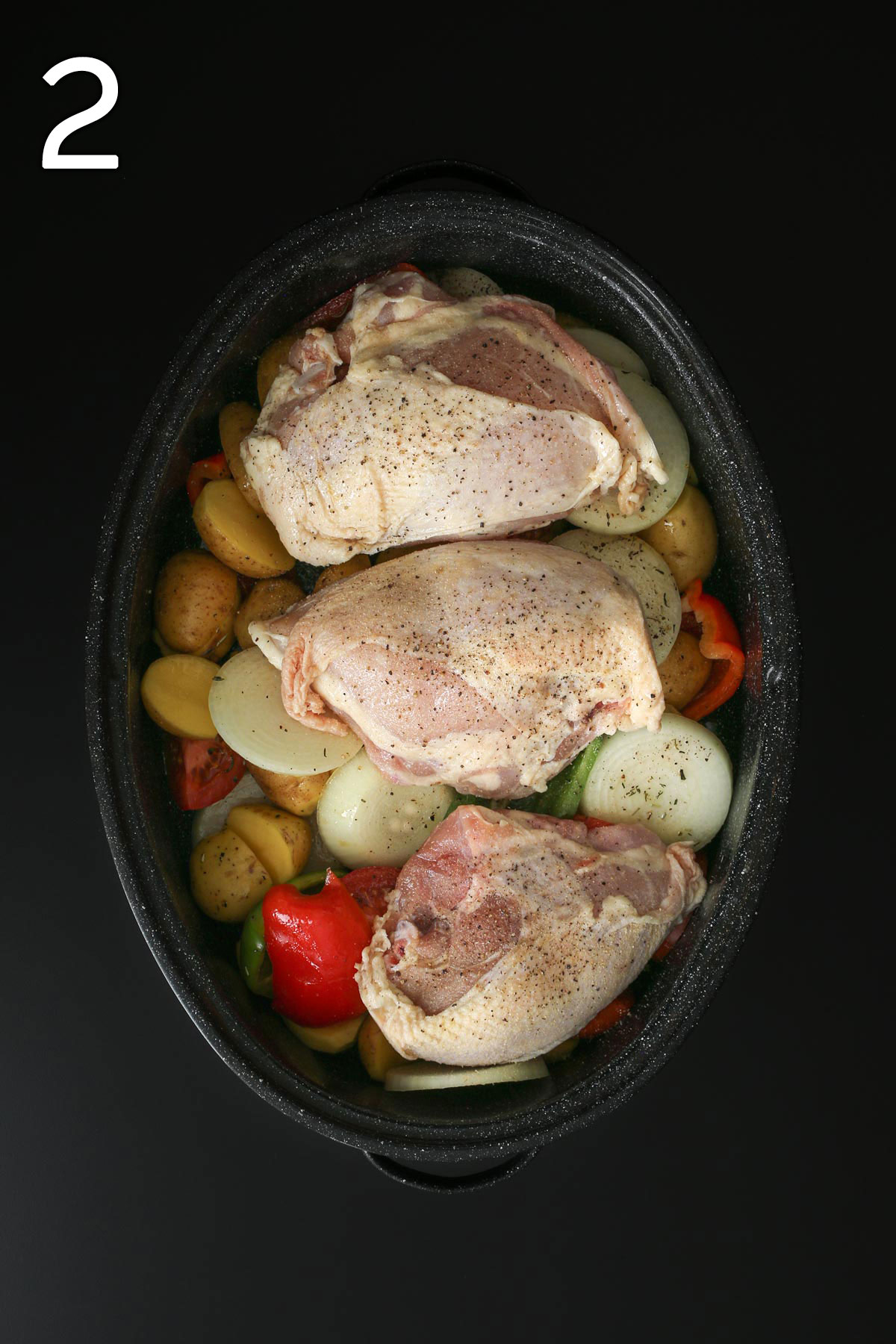 uncooked chicken pieces atop the vegetable mixture in the roasting pan on black table top.