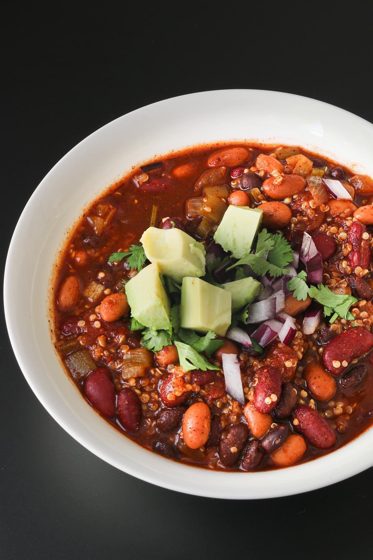 close up of a white bowl of quinoa chili topped with chopped cilantro, cubed avocado, and chopped red onion.