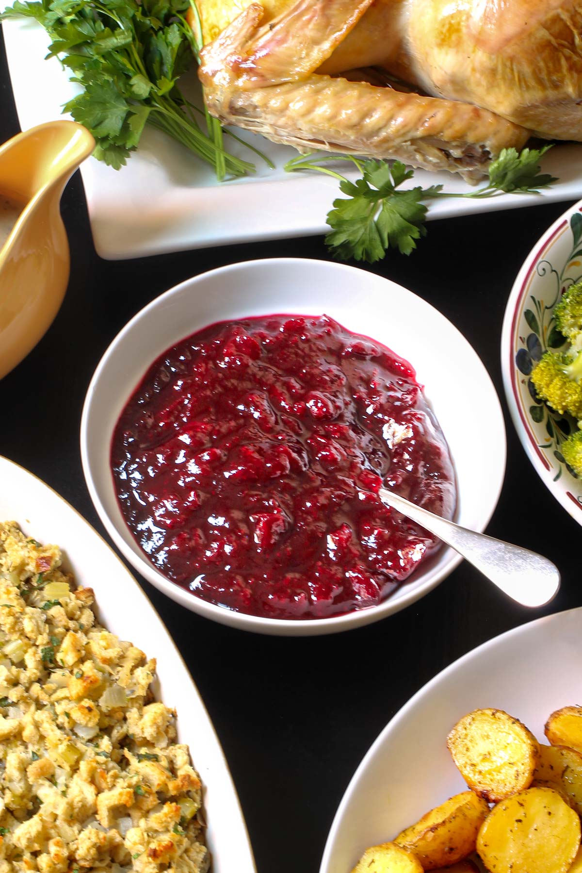 a white bowl of cranberry sauce with a spoon on a table set with other dinner platters.