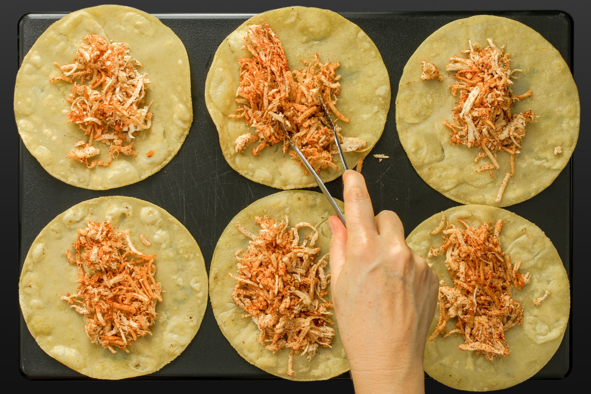 using tongs to lay cooked and seasoned shredded chicken on fried tortillas for enchiladas or flautas.