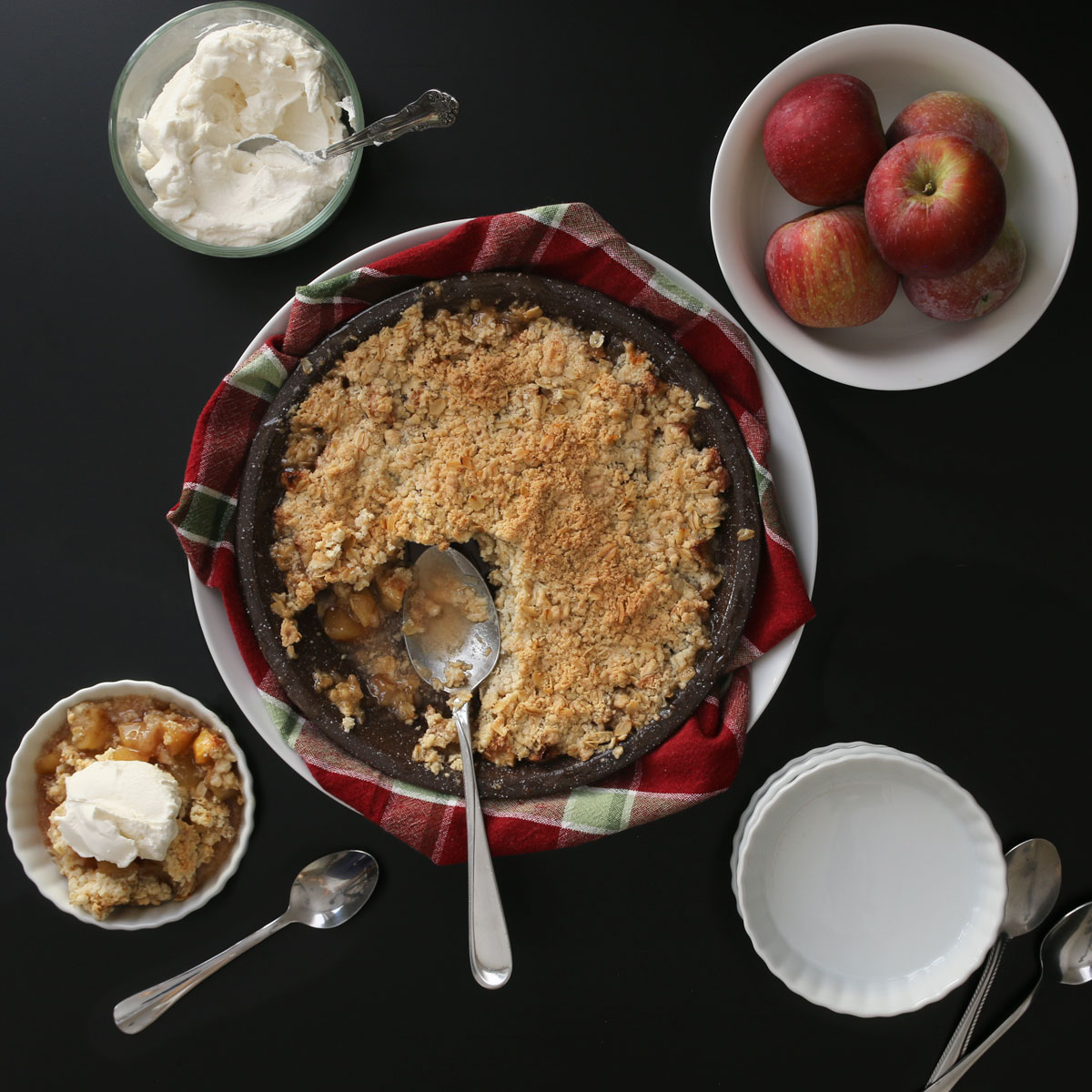 pan of apple crisp surrounded by whole apples, a bowl of whipped cream, and plates for dishing up.