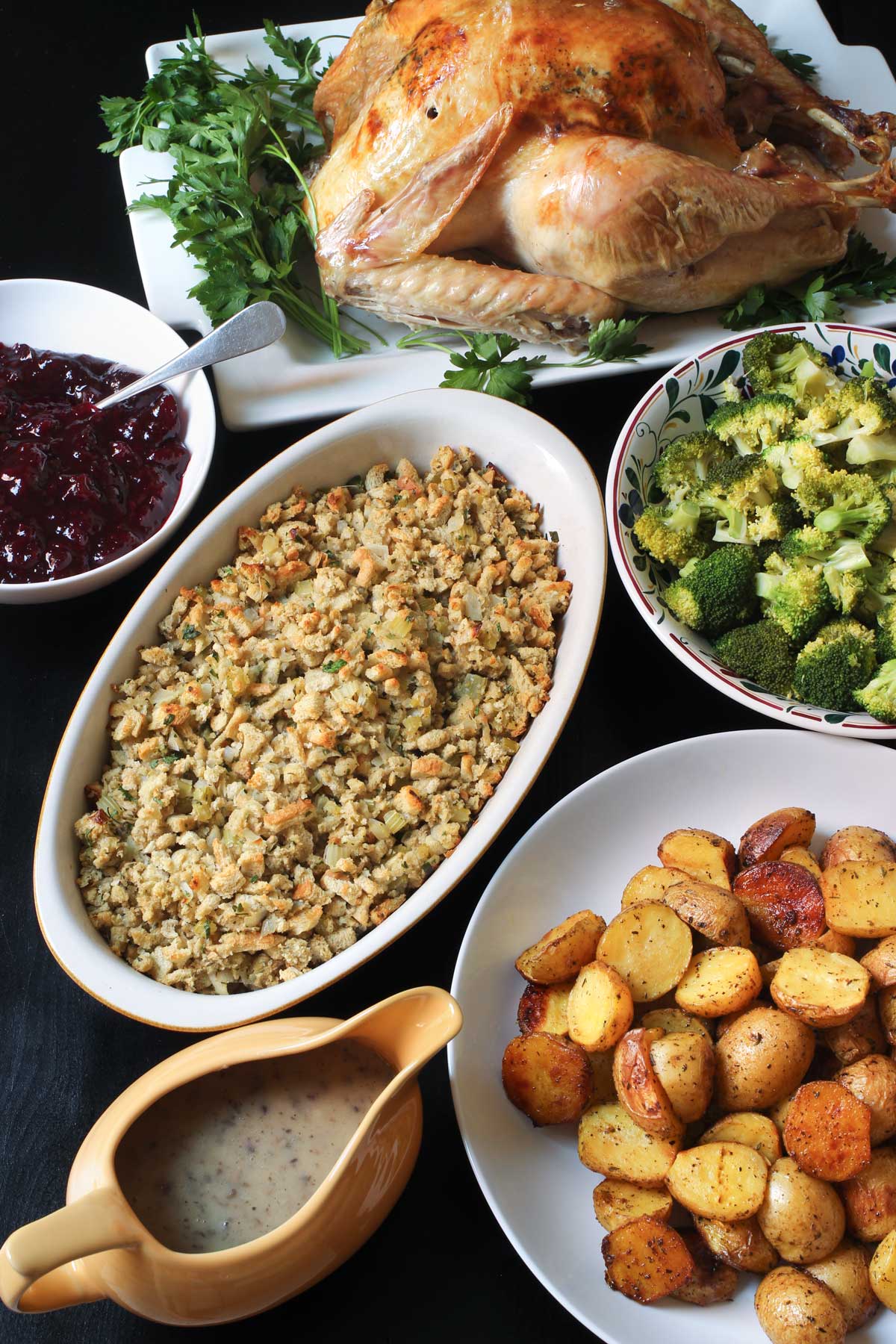 thanksgiving dinner laid out table: roast turkey, broccoli, stuffing, cranberry sauce, gravy, and roast potatoes.