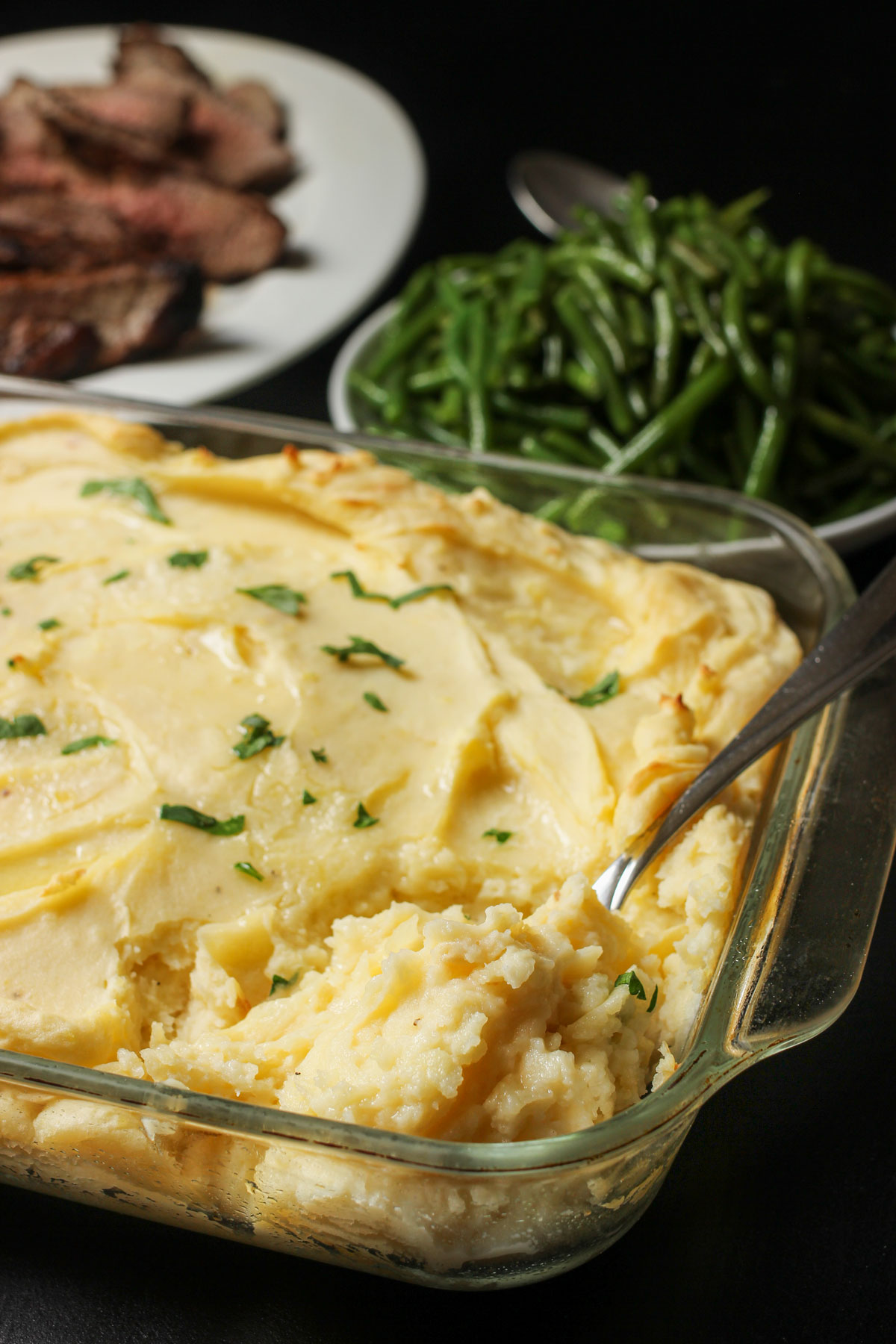baked mashed potato casserole topped with chopped fresh parsley on dinner table next to bowl of green beans and a platter of sliced tri-tip.