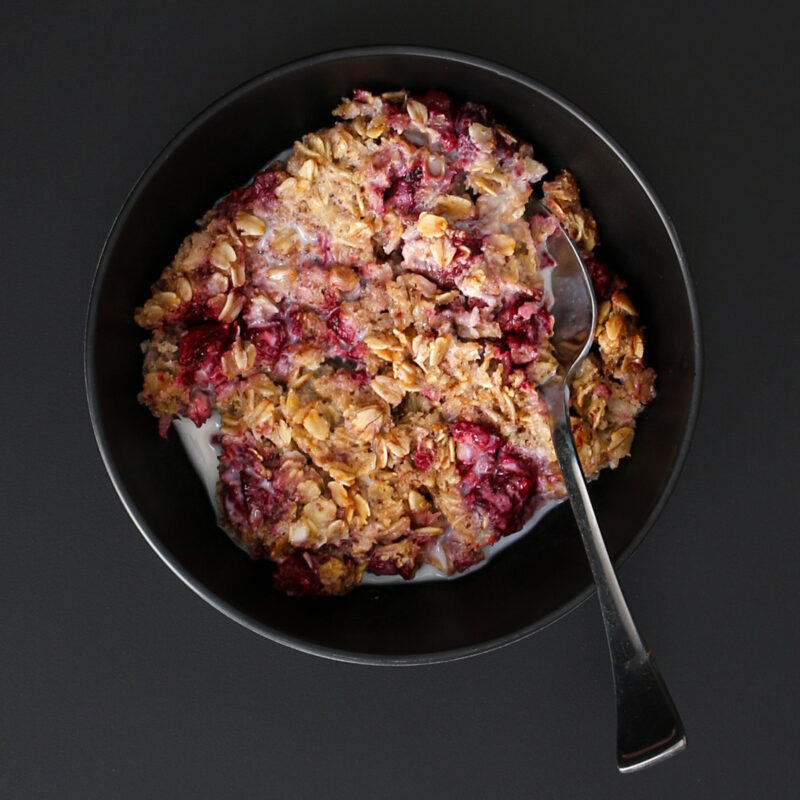 overhead shot of black bowl of vegan baked oatmeal with raspberries and almond milk with a spoon immersed.