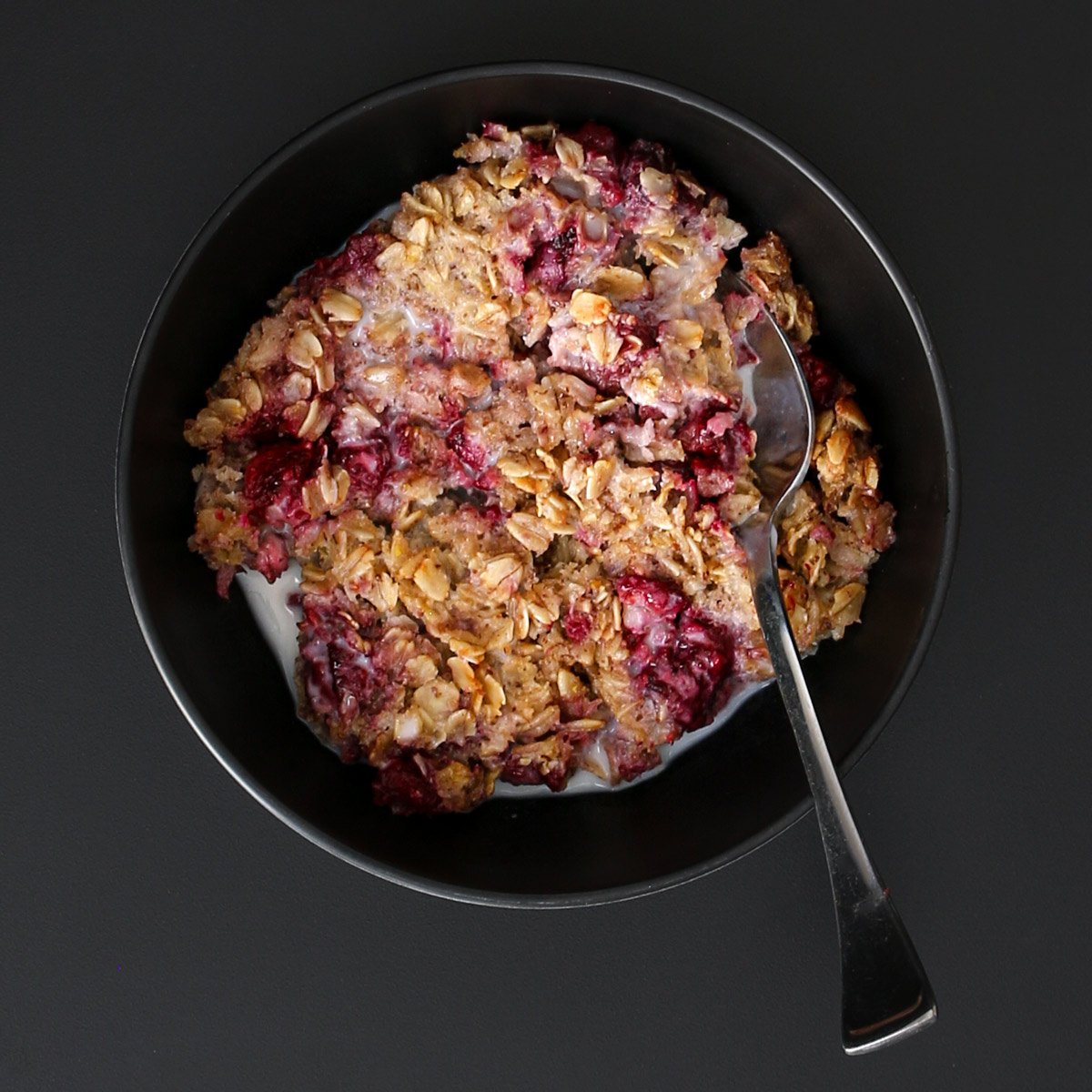 overhead shot of black bowl of vegan baked oatmeal with raspberries and almond milk with a spoon immersed.