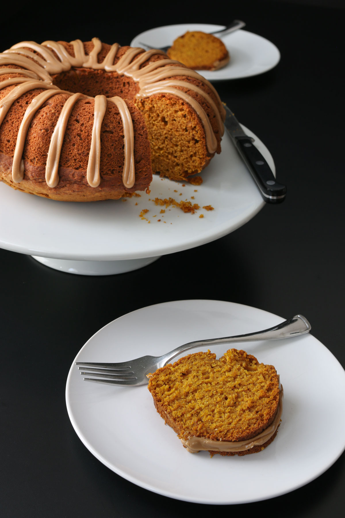 pumpkin bundt cake on white stand on black table with plates of cake with forks nearby.