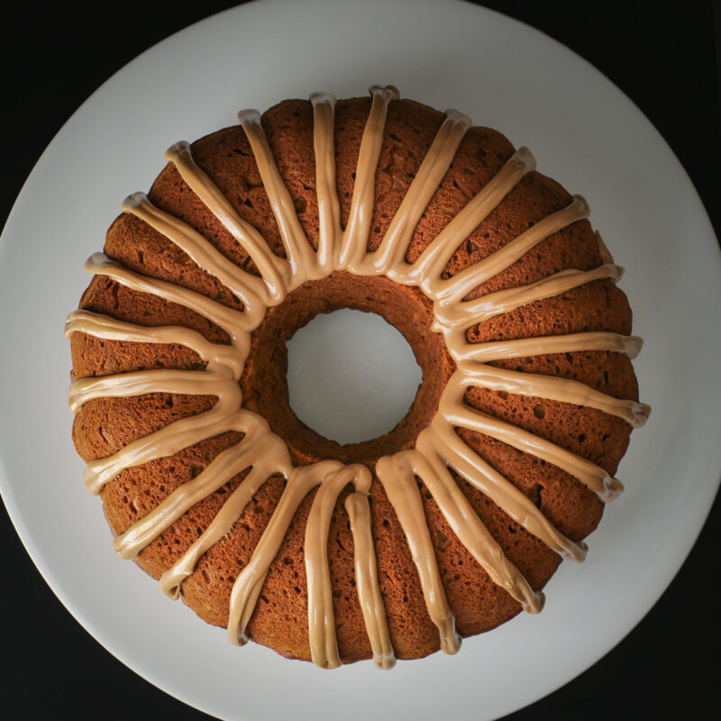 overhead shot of pumpkin bundt cake with maple glaze piped in stripes on white platter on black table.